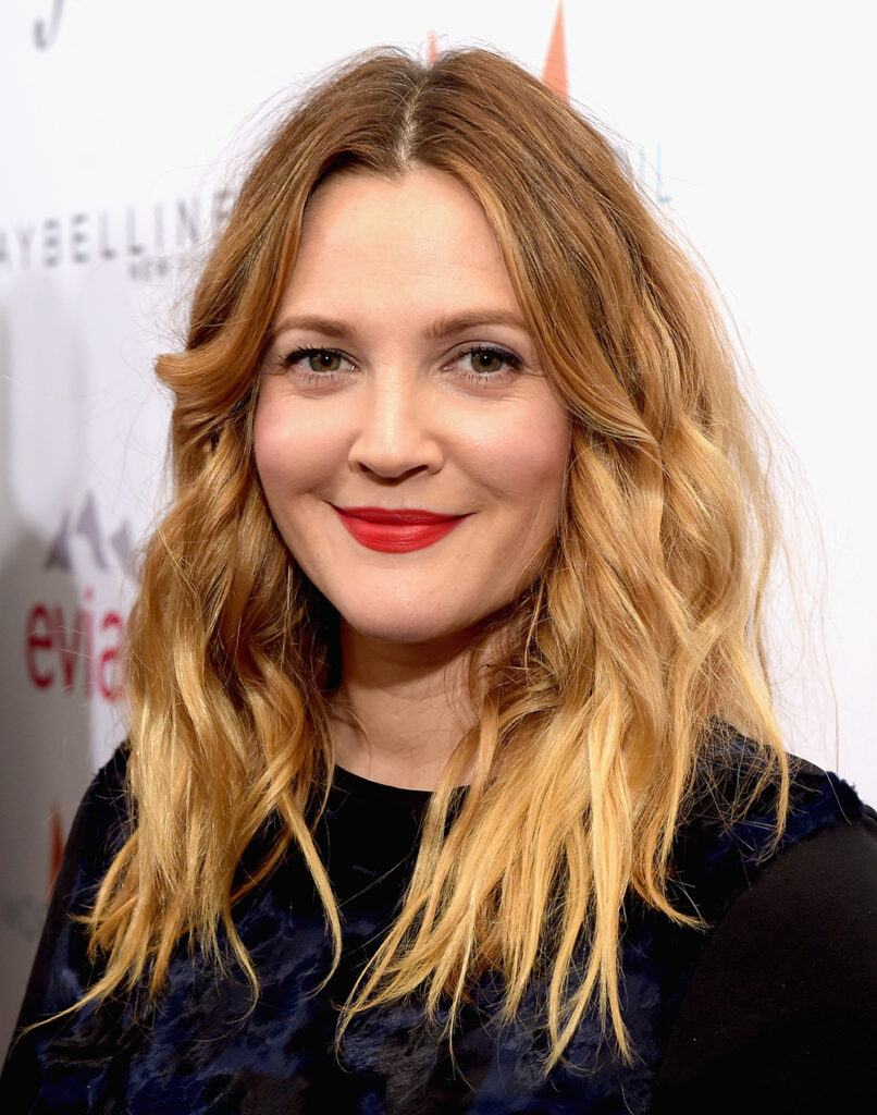 Actress Drew Barrymore wearing a black dress with red lipstick, blonde wavy hair.